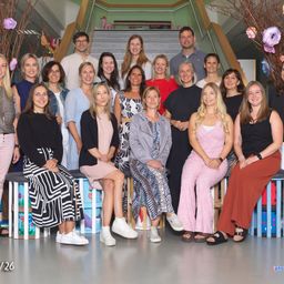 Eine Gruppe von 26 Menschen, darunter Männer und Frauen, posiert für ein Foto vor einer Treppe. Sie tragen alle Freizeitkleidung und einige von ihnen tragen eine Brille. Die Frau in der Mitte hält eine Handtasche.