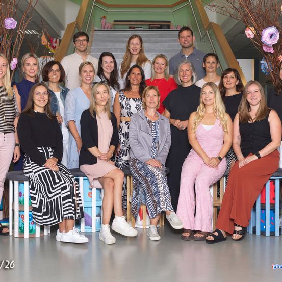 Eine Gruppe von 26 Menschen, darunter Männer und Frauen, posiert für ein Foto vor einer Treppe. Sie tragen alle Freizeitkleidung und einige von ihnen tragen eine Brille. Die Frau in der Mitte hält eine Handtasche.