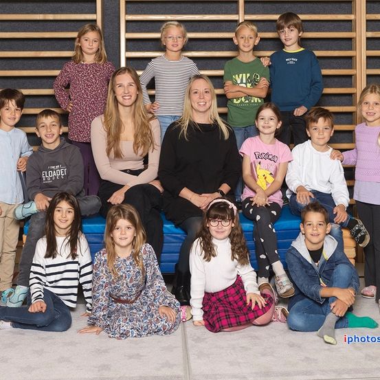 Eine Gruppe von Kindern und zwei Frauen posiert für ein Foto, stehend und sitzend auf einem blauen Kissen. Sie befinden sich in einem Raum mit einer Holzwand und einem Teppichboden.