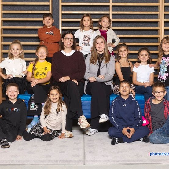 Eine Gruppe von Kindern und zwei Frauen posiert für ein Foto in einem Fitnessstudio. Die Kinder sitzen und stehen auf einer blauen Bank und lächeln für die Kamera. Die Frauen sitzen in der Mitte der Bank. Das Bild hat einen Wasserzeichen auf der unteren rechten Seite.