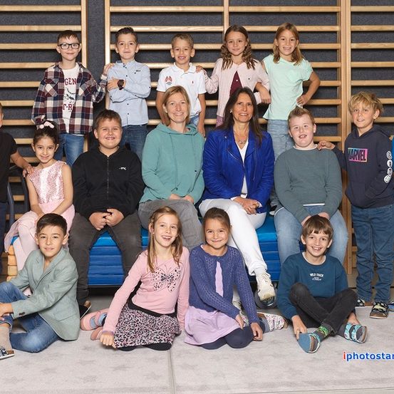 Eine Gruppe von Kindern und zwei Frauen posiert für ein Foto vor einer Holzwand. Die Kinder sitzen auf dem Boden und einer Bank.