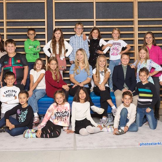 Eine Gruppe von Kindern und zwei Erwachsenen posiert für ein Foto. Sie sitzen und stehen auf einem Teppichboden. Einige tragen Brillen. Sie befinden sich in einem Studio mit Holzstäben dahinter.