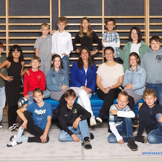 Eine Gruppe von Kindern und Erwachsenen posiert für ein Foto vor einer Holzwand. Die Gruppe umfasst eine Frau in einer blauen Jacke.