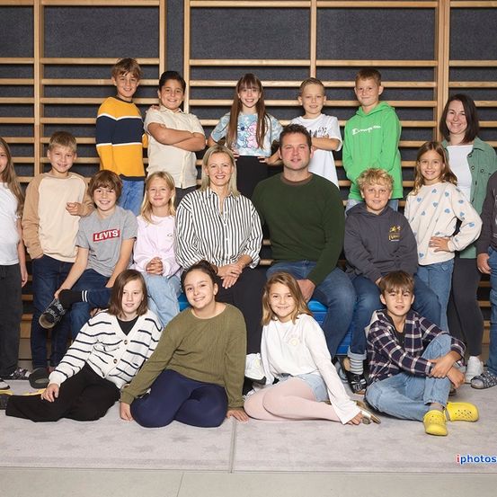 Eine Gruppe junger Menschen und zwei Erwachsene posieren für ein Foto vor einer Holzwand. Einige sitzen auf dem Boden, andere stehen.