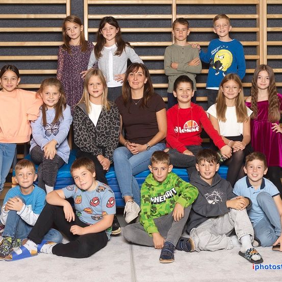 Ein Gruppenfoto von Kindern und zwei Erwachsenen, alle lächelnd und posierend vor einer Holzwand. Einige sitzen auf einer blauen Bank.