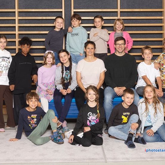Eine Gruppe von Kindern und Erwachsenen posiert für ein Foto, einige sitzen auf einer blauen Bank und andere stehen dahinter. Eine Frau mit Brille und ein Mann in einem schwarzen Shirt sind in der Mitte.