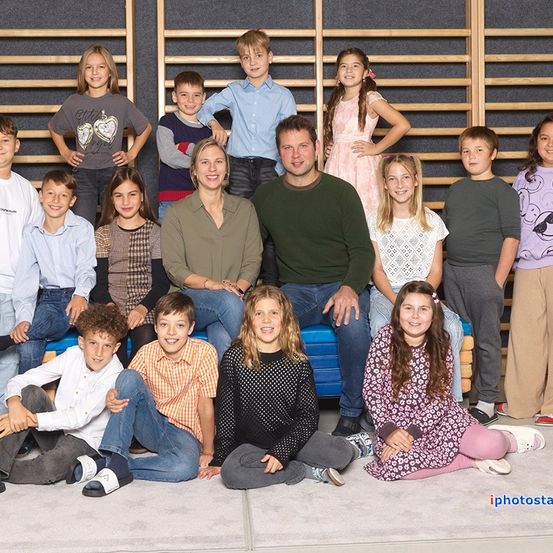 Eine Gruppe von Kindern und Erwachsenen posiert für ein Foto. Sie sitzen und stehen auf einer blauen Bank vor einer Holzwand. Einige von ihnen lächeln. Das Bild hat einen Wasserzeichen auf der unteren rechten Seite.