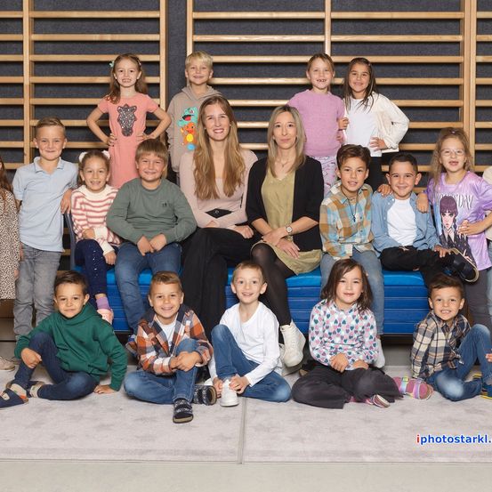Eine Gruppe von Kindern und zwei Frauen posiert für ein Foto. Die Kinder sitzen und stehen in zwei Reihen, mit einer blauen Bank in der Mitte. Die beiden Frauen stehen hinter der Bank. Die Kinder tragen verschiedene Kleidungsstücke und Schuhe. Die Holzwand hinter ihnen hat ein Design.