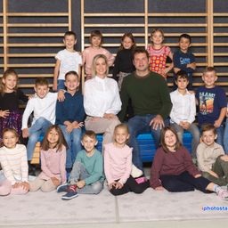 Eine Gruppe von Kindern und zwei Erwachsenen posiert für ein Foto im Klassenzimmer. Die Kinder sitzen und stehen auf einem blauen Kissen.