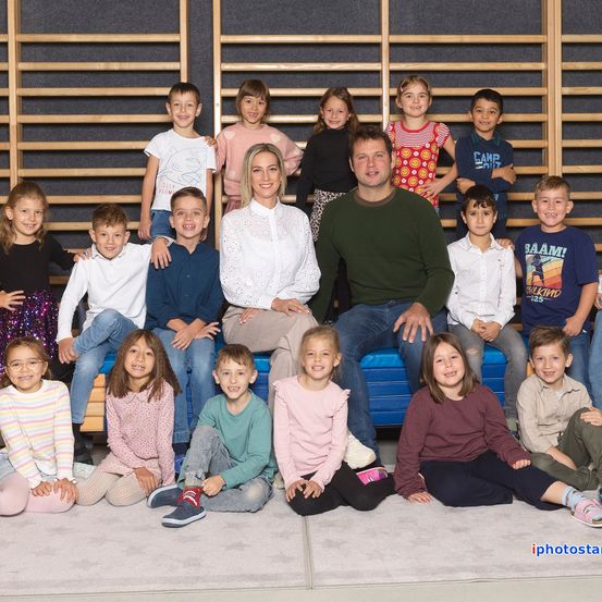 Eine Gruppe von Kindern und zwei Erwachsenen posiert für ein Foto im Klassenzimmer. Die Kinder sitzen und stehen auf einem blauen Kissen.