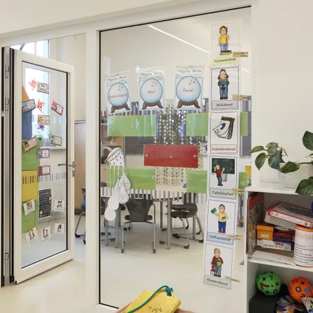 Ein Klassenzimmer mit einer Glastür, farbenfrohen Dekorationen und verschiedenen Postern. Ein Tisch mit Stühlen spiegelt sich im Glas. Eine Topfpflanze steht auf einem Regal mit Spielzeug.