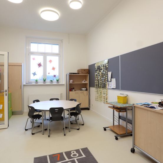 Ein leerer Klassenraum mit einem runden Tisch und Stühlen darum herum. Der Raum hat weiße Wände und eine weiße Decke mit kreisförmigen Lichtern. Ein Schrank mit Regalen steht neben dem Tisch. Ein Fenster mit Topfpflanzen ist an der Wand.