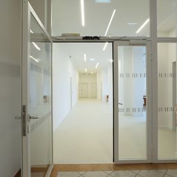 Ein leerer Flur mit weißen Wänden und Böden, zwei Türen am Ende und Glaswänden mit einem Datenschutzmuster.