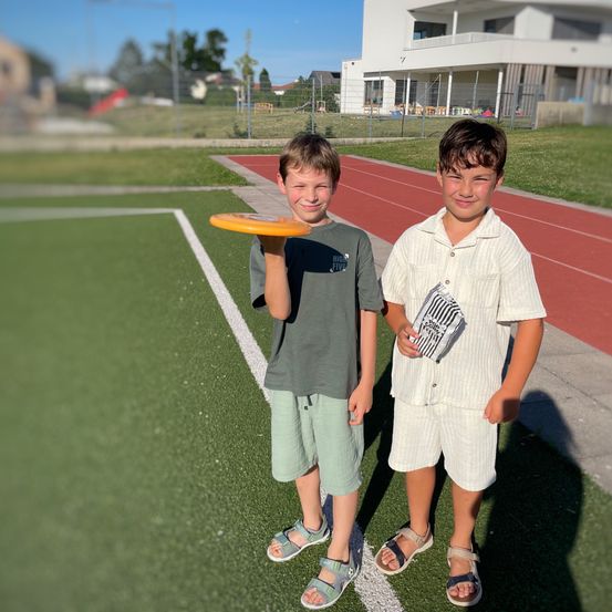 Zwei Jungen stehen auf einem Sportplatz mit einem Frisbee. Ein Junge hält eine Tasche. Hinter ihnen steht ein Gebäude und ein Spielplatz.