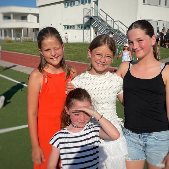 Vier Mädchen posieren für ein Foto auf einem Sportplatz, eines bedeckt sich die Augen mit der Hand. Dahinter befindet sich ein Gebäude mit einer Treppe und einem Rasenbereich mit Menschen.