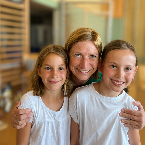 Eine lächelnde Frau mit zwei jungen Mädchen, alle in weißen T-Shirts, stehen gemeinsam in einem Raum mit Holzwänden.