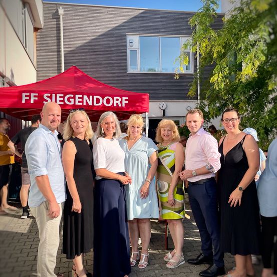 Eine Gruppe von Menschen, darunter drei Frauen und drei Männer, steht unter einem roten Zelt und lächelt für ein Foto. Sie sind lässig gekleidet, und das Zelt steht 'FF SIEGENDORFF'. Hinter ihnen befindet sich ein Gebäude mit Fenstern.