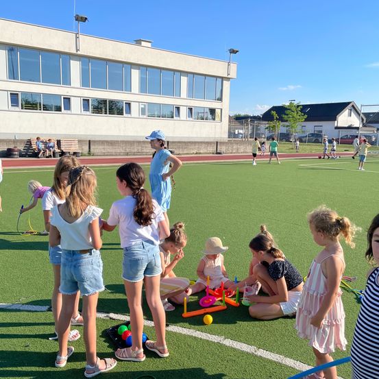 Eine Gruppe von Kindern spielt auf einem Freiluftfeld. Einige sitzen auf dem Boden, während andere stehen und zusehen. Das Feld hat ein Gebäude mit Glasfenstern im Hintergrund.