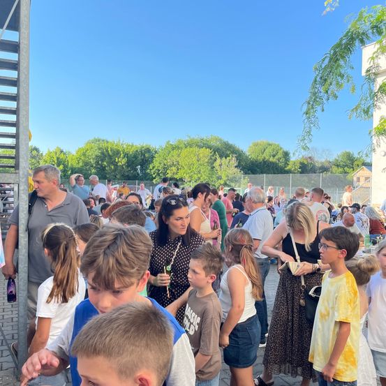 Eine Menschenmenge aus Erwachsenen und Kindern versammelt sich tagsüber in einem Außenbereich, wahrscheinlich für eine Veranstaltung. Einige stehen, andere gehen. Bäume und ein Zaun sind im Hintergrund.