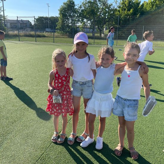 Vier junge Mädchen stehen auf einem Sportplatz, lächeln und posieren für ein Foto. Sie tragen lässige Sommerkleidung. Hinter ihnen bilden ein Zaun und Bäume eine Kulisse. In der Ferne sind andere Kinder zu sehen.