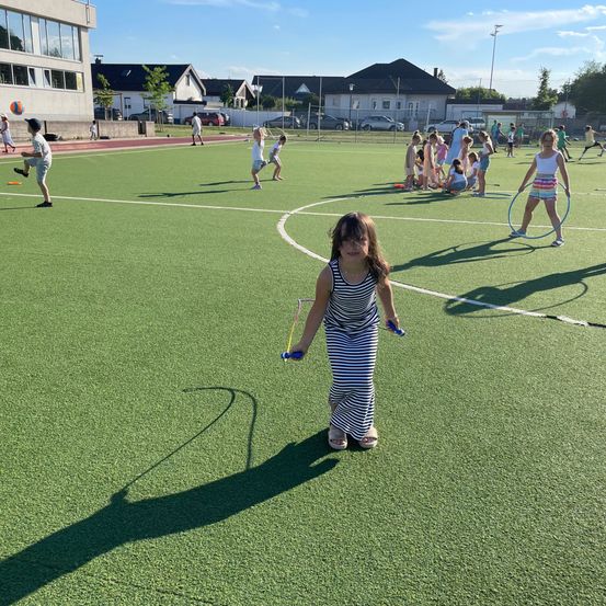 Kinder spielen mit Hula-Hoops auf einem grünen Feld mit verschiedenen Gebäuden im Hintergrund unter einem bewölkten Himmel.