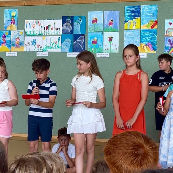 Mehrere Kinder stehen vor einer grünen Wand mit Gemälden. Sie halten rote Papiere und einer ein Mikrofon. Sie sehen ernst aus. Hinter ihnen sitzt ein Junge auf dem Boden.