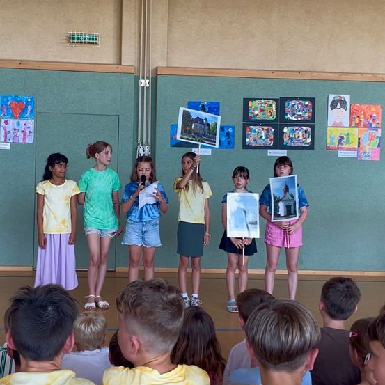 Mehrere junge Mädchen stehen auf der Bühne und halten Bilder in den Händen, eines der Mädchen hält ein Mikrofon. Dahinter zeigt eine grüne Wand verschiedene Kunstwerke. Davor sitzen mehrere Kinder und beobachten die Präsentation.