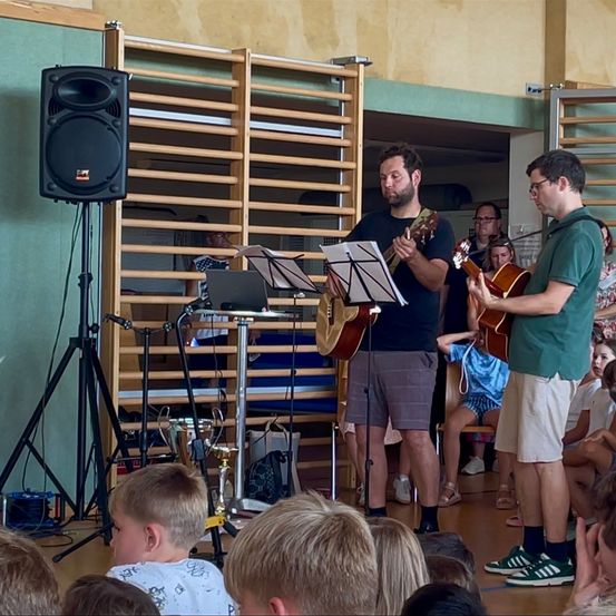 Zwei Männer spielen Gitarren, während sie vor einem kleinen Publikum stehen, möglicherweise in einem Klassenzimmer. Der Mann rechts trägt eine Brille, und der Mann links trägt Turnschuhe. Hinter ihnen steht ein Lautsprecher, ein Notenständer und andere Musikinstrumente.