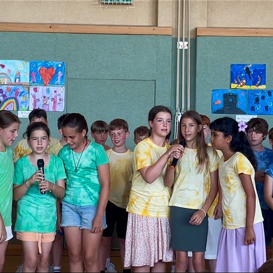 Eine Gruppe von Kindern steht in einer Reihe und trägt gelbe und grüne T-Shirts. Einige halten Mikrofone, was darauf hindeutet, dass sie gleich auftreten. Der Raum hat farbenfrohe Kunst an der Wand.