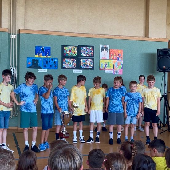 Eine Gruppe von Jungen in blauen und gelben T-Shirts steht in einer Reihe und hält einen Pokal, mit Gemälden an der Wand hinter ihnen und einem Lautsprecher rechts.