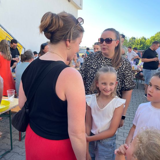 Eine Gruppe von Erwachsenen und Kindern, darunter zwei junge Mädchen, stehen zusammen draußen. Die Erwachsenen unterhalten sich. Im Hintergrund sind Tische aufgebaut, und Menschen sitzen und stehen.