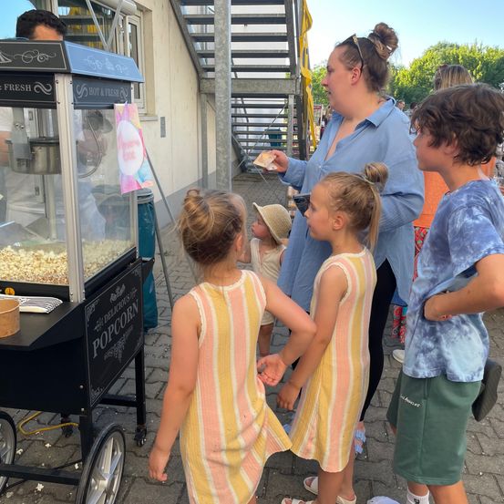Eine Frau hält Geld in der Hand und steht neben einer Popcornmaschine. Zwei junge Mädchen stehen vor ihr, während ein Junge zusieht. Ein Baby in einem Hut steht hinter ihnen.