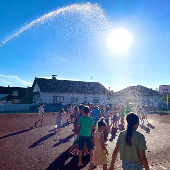 Eine Gruppe von Kindern spielt an einem sonnigen Tag unter einem Wassersprüher, Schatten auf dem Boden, in der Nähe von Häusern und einem Basketballplatz.