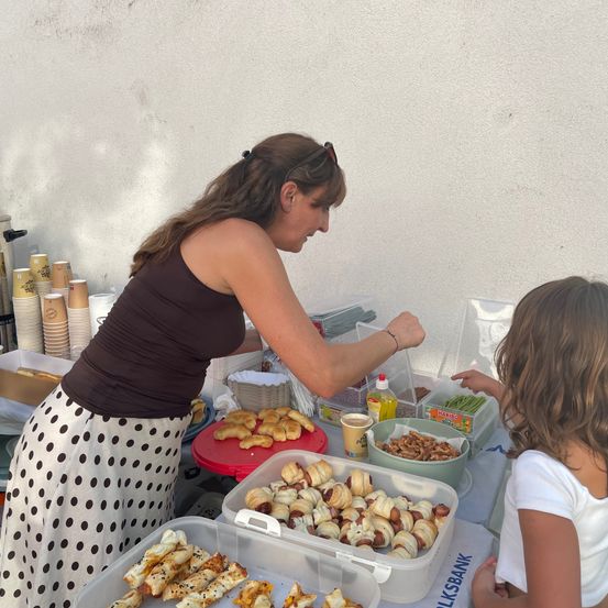 Eine Frau serviert einer kleinen Mädchen an einem Tisch mit verschiedenen Snacks und Getränken bei sonnigem Wetter.