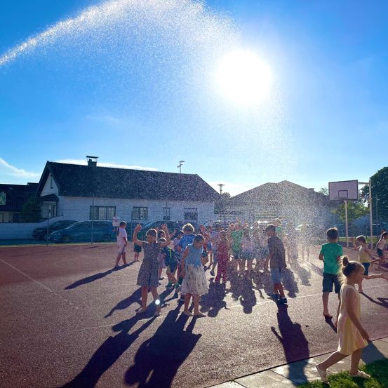 Eine Gruppe von Kindern spielt und spritzt unter einem Sprinkler an einem sonnigen Tag auf einem Freiluftsportplatz.