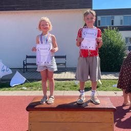 Zwei Kinder stehen auf einer Holzkiste mit Urkunden in der Hand, eines trägt eine Medaille. Hinter ihnen steht ein Gebäude mit Glasfenstern, einer Bank und einer Flagge.