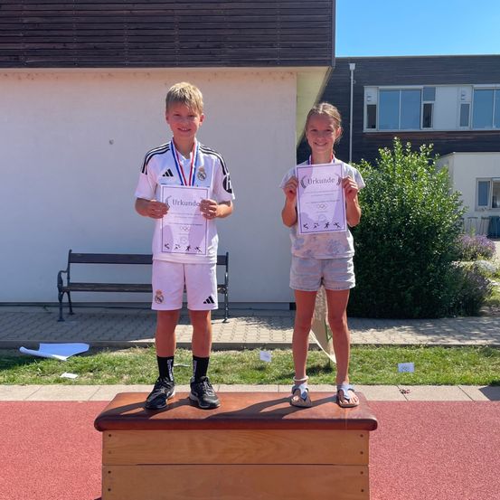 Zwei Kinder stehen auf einem Podest, halten Urkunden und Medaillen. Der Junge trägt eine Medaille um den Hals. Sie lächeln beide. Hinter ihnen steht eine Bank und ein Gebäude.