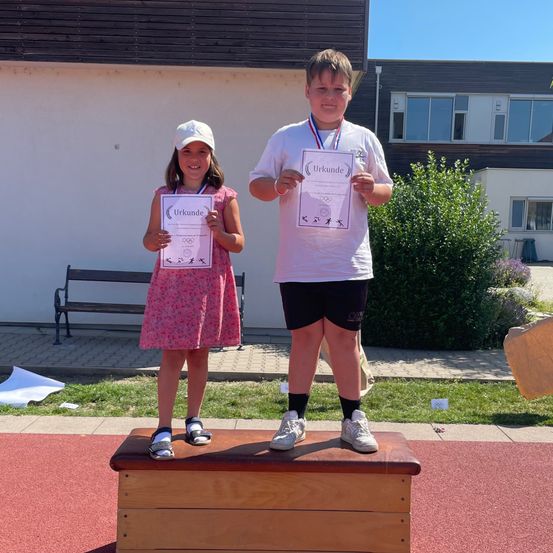 Zwei Kinder stehen auf einem Podest und halten Urkunden und Medaillen in den Händen. Das Mädchen trägt ein rosa Kleid, und der Junge ein weißes T-Shirt und schwarze Shorts. Hinter ihnen steht ein Gebäude mit Glasfenstern und einer Bank.
