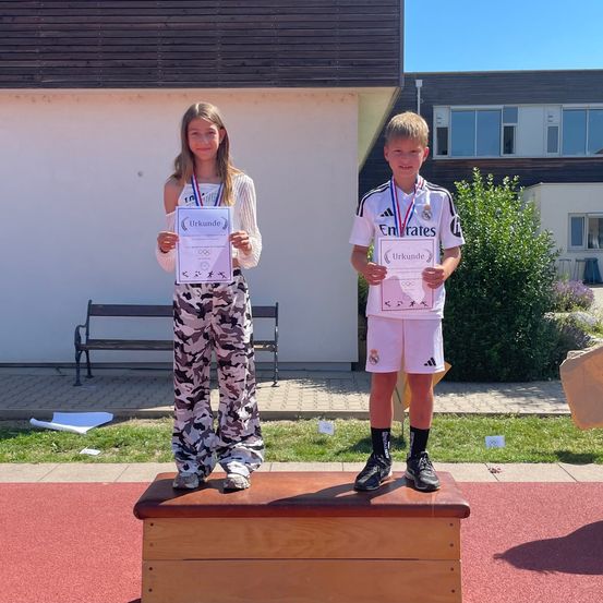 Zwei Kinder stehen mit Medaillen und Urkunden in den Händen auf einem Podium. Sie sind draußen vor einem Gebäude mit einer Bank und einigen Pflanzen.