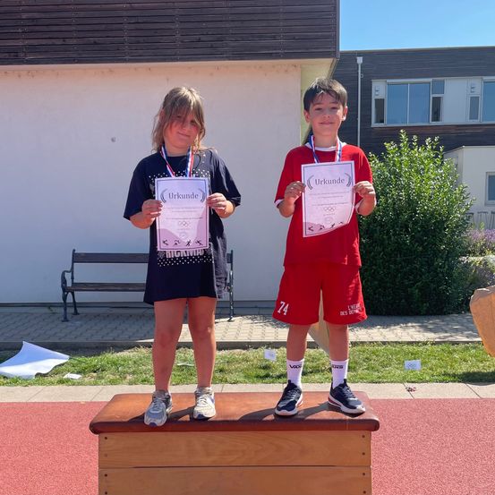 Zwei Kinder stehen auf einem Podium und halten Urkunden in den Händen. Ein Mädchen in einem schwarzen Shirt und ein Junge in einem roten Shirt, beide lächeln. Hinter ihnen steht ein Gebäude, eine Bank und ein Baum.