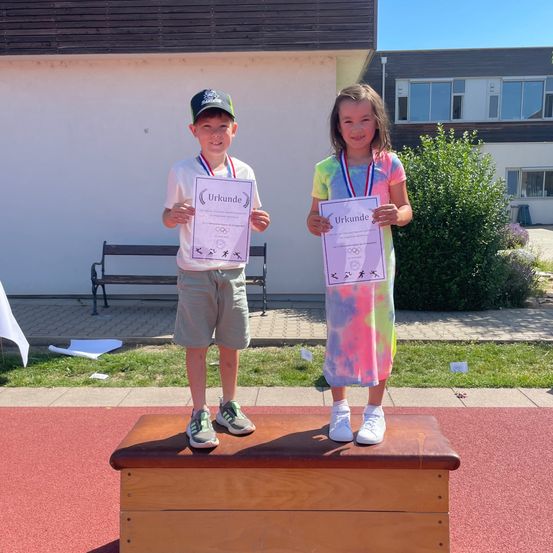 Zwei Kinder stehen auf einer Holzkiste und halten Urkunden in den Händen. Der Junge trägt eine Baseballkappe, Turnschuhe und eine Medaille. Das Mädchen ist in einem Tie-Dye-Kleid, ebenfalls mit einer Medaille. Dahinter steht eine Bank, Pflanzen und ein Gebäude.