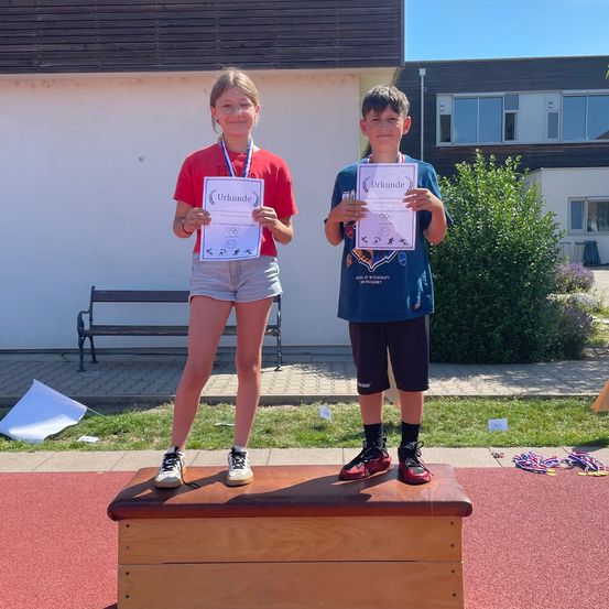 Zwei Kinder stehen auf einem Podium, halten Urkunden in der Hand, mit Medaillen und einer Bank im Hintergrund.