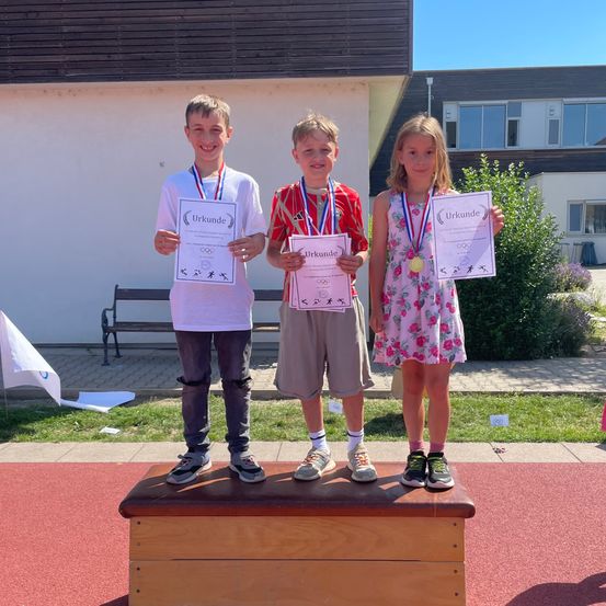 Drei Kinder stehen auf einem Podest und halten Urkunden in der Hand. Zwei Jungen und ein Mädchen, alle mit Medaillen. Sie sind draußen auf einer roten Laufbahn. Dahinter ein weißes Gebäude mit Glasfenstern.