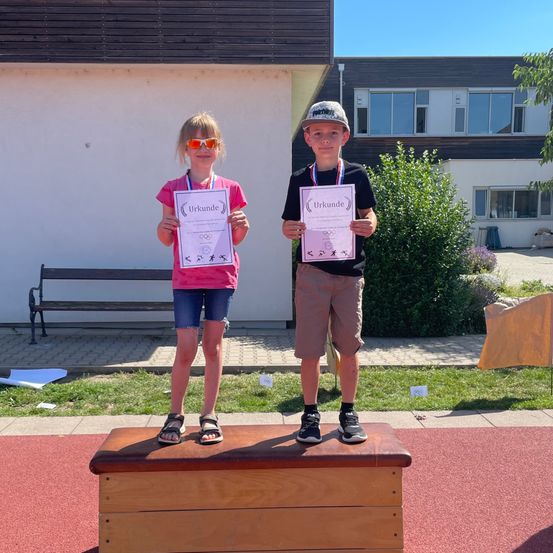 Zwei kleine Kinder stehen auf einer Holzkiste und halten Urkunden in den Händen. Sie tragen Medaillen um den Hals und Sonnenbrillen. Sie sind auf einer roten Laufbahn vor einem Gebäude mit einer Bank und einem Busch.