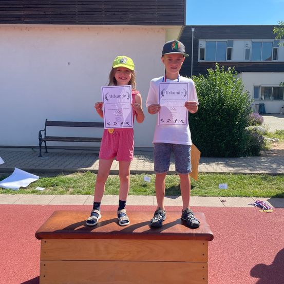 Zwei Kinder stehen auf einem Holzkasten und halten Urkunden in den Händen. Das Mädchen lächelt und der Junge schaut in die Kamera. Hinter ihnen steht ein Gebäude mit Glasfenstern und einer Bank.