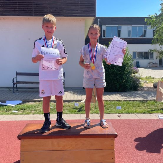Zwei Kinder stehen auf einem Podium mit Medaillen um den Hals und Urkunden in den Händen. Der Junge trägt ein weißes Shirt mit Logo, während das Mädchen ein weißes Oberteil mit Blumenmuster trägt. Dahinter steht ein Gebäude mit Fenstern und eine Bank auf der linken Seite.