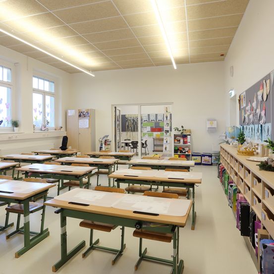 Ein leeres Klassenzimmer mit weißen Wänden und weißem Boden. Es hat Schreibtische und Stühle und eine Ankündigungstafel auf der rechten Seite. Links befinden sich Fenster mit Topfpflanzen und Zeichnungen.