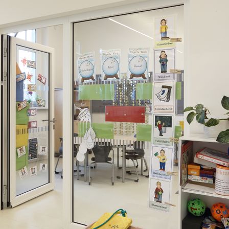 Ein Klassenzimmer mit Glastür und Fenstern. Drinnen stehen Stühle und ein Schreibtisch, zusammen mit Postern und Dekorationen. Rechts steht eine Topfpflanze und unten ein Regal mit Spielzeug.