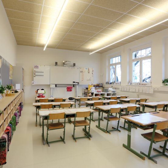 Ein leerer Klassenraum mit weißen Wänden und Decke. Reihen von Schreibtischen und Stühlen sind vor einer Tafel angeordnet. Auf der rechten Seite befinden sich drei Fenster und ein Regal mit Büchern und Pflanzen auf der linken Seite.