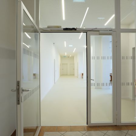 Ein leerer Korridor mit weißen Wänden, gefliestem Boden und zwei Glastüren. Eine Reihe von Lichtern verläuft entlang der Decke.